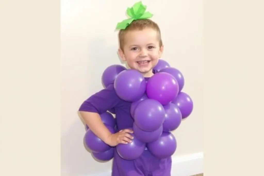 Dziecko w kostiumie winogron wykonanym z fioletowych balonów przyczepionych do ubrania